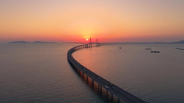 Aerial parallax of Incheon Bridge curving over calm sea with traffic silhouette against blazing orange sunset and cargo ships