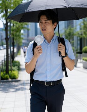 夏のオフィス街で日傘とハンディファンを使い暑さ対策をする若い男性会社員