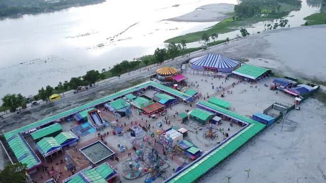 Drone shot of a busy riverside funfair with ferris wheel slides stalls people bordered by a river and a distant bridge under a cloudy sky carnival amusement park rides landscape aerial