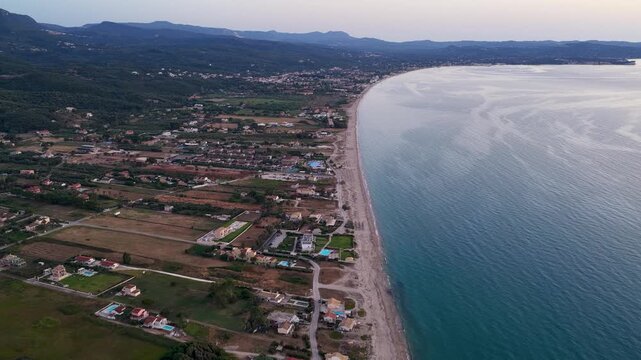A stunning high-altitude aerial drone shot capturing the expansive sandy coastline, calm Ionian Sea, and seaside developments of Almyros and Acharavi on Corfu Island, Greece, during a peaceful evening