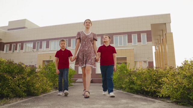 Two boys and a woman walking outside, They walk in front of a new building, The boys hold the woman&rsquo;s hands, Their heads are up, The boys are in matching outfits, The woman has a floral dress, The