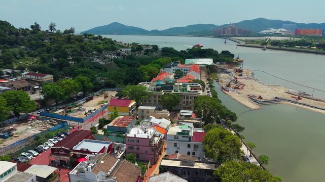 Aerial pullback rises above Coloane Village rooftops revealing sea channel with passing ferry boat and Cotai resort skyline on horizon