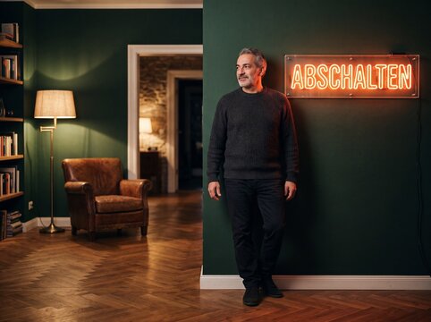 Reifer Mann lehnt l&auml;chelnd an gr&uuml;ner Wand neben leuchtendem Neonschild mit Wort Abschalten im gem&uuml;tlichen Wohnzimmer