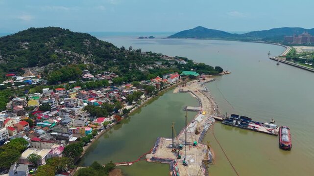 Aerial view ofl Coloane Village rooftops beside canal and coastal land reclamation site with green hills and sea in distance