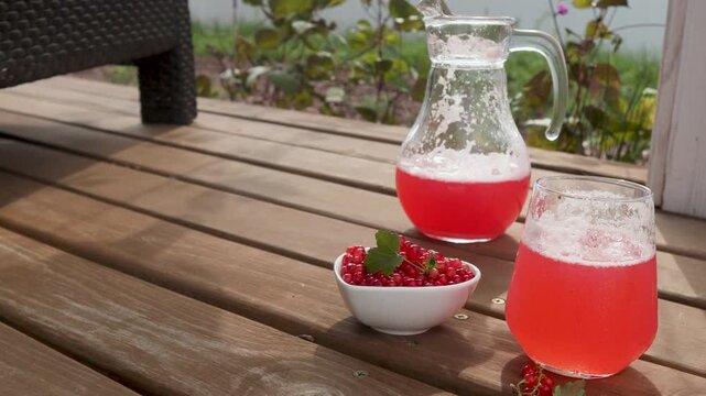 Wooden patio table holds glass jug and glass of red currant drink beside small bowl of fresh berries, framed by garden furniture and greenery in warm sunlight.