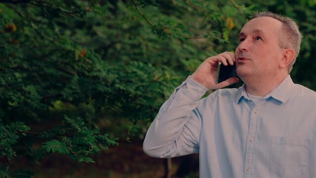 casual attire, casual blue shirt, listening intently happily., smiling caller in nature, standing outside amid lush natural surroundings., standing by leafy branches nearby., happy middle-aged man
