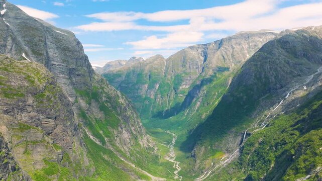 Majestic panoramic view of a vast green valley flanked by towering rocky mountains with dramatic waterfalls cascading down steep slopes under a bright blue sky with scattered clouds