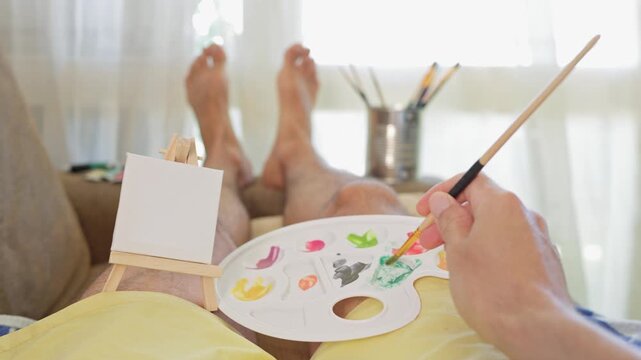 point of view of an artist relaxing on a sofa while mixing green paint on a palette before starting to paint on a tiny canvas supported by a miniature easel, a creative and relaxing hobby