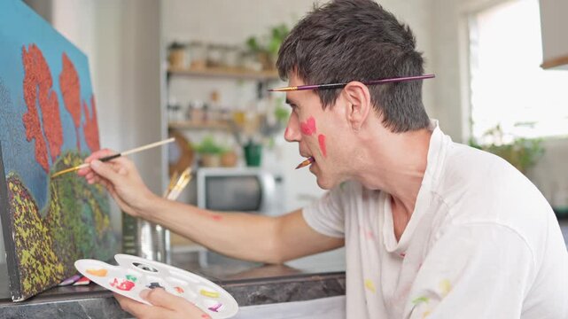 Passionate artist with a messy face and paint-stained clothes focusing intently on creating a masterpiece, holding a palette and brush while working on a colorful canvas in his home studio