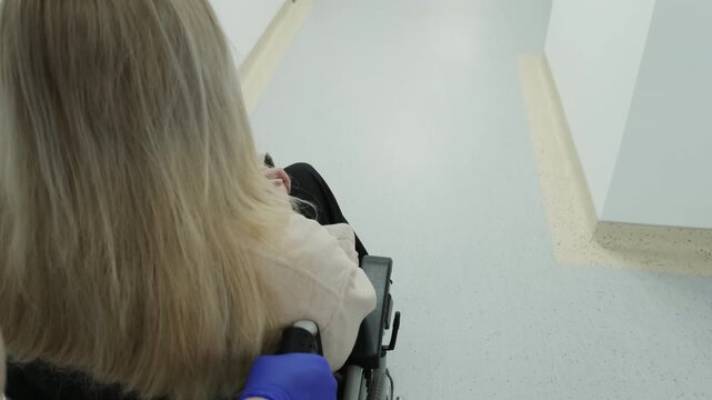 Nurse helps disabled woman patient reach ultrasound examination room. Doctor pushes wheelchair through hospital corridor. Slow motion