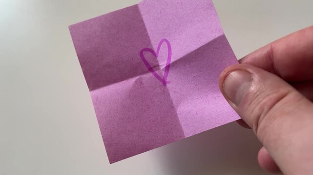 Hand Placing Pink Folded Note with Drawn Heart Symbol on White Surface - Valentine's Day Love Concept