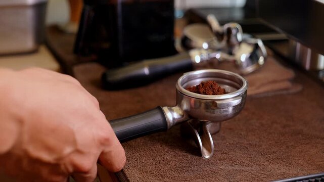 Close-up of a barista's hands pressing ground coffee with a wooden-handle tamper into a stainless steel portafilter, preparing espresso shot for a professional coffee machine.	