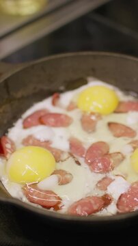 Vertical video. Hot breakfast pan. Frying egg. Pork sausage slices roasting in boiling bubbling steaming oil. Cooking tasty meal on black electric stove.
