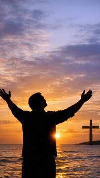 A man praying with his arms raised towards a beautiful sunset over the ocean.