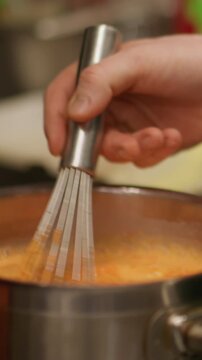 Vertical video. Cooking process. Chef whisking. Metal saucepan orange liquid heating on stove. Hand mixing soup ingredients for soft delicious dish.