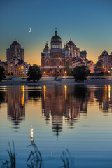Fototapeta premium Young crescent moon setting after sunset above the Cathedral of the Nativity of Christ on Obolon Embankment in reflected in the calm waters of the Dnipro River