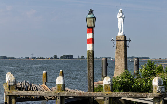 Volendam, Netherlands - White statue of the Virgin Mary standing on a stone pedestal beside a red and white harbor marker pole at the entrance of Volendam harbor.