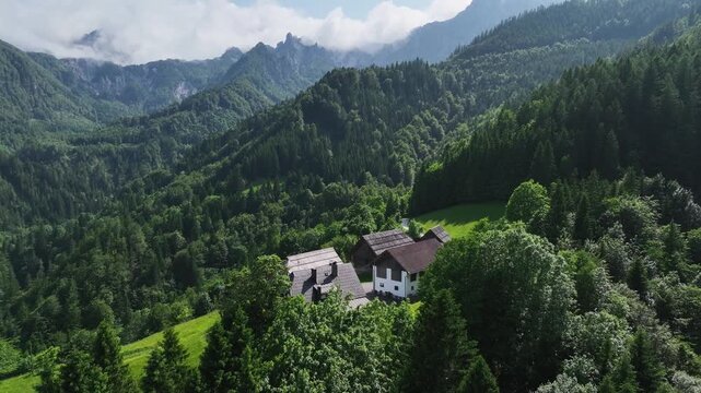 Drone captures winding asphalt road crossing lush green mountain valley in Austria. Traditional houses sit near forest edge during sunny summer day in Alpine region near border.