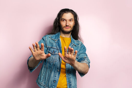 Young man with long hair in denim jacket doing stay away gesture. On pastel pink background.