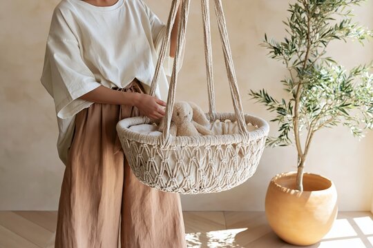 Young woman in oversized cotton t-shirt and wide-leg trousers placing plush bunny in woven macrame cradle in a boho nursery setting