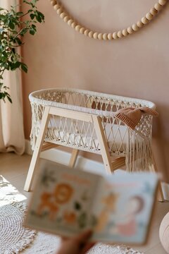 Illustrated baby board book held open in the foreground, focusing on a woven macrame cradle with a knit hat and wooden bead garland