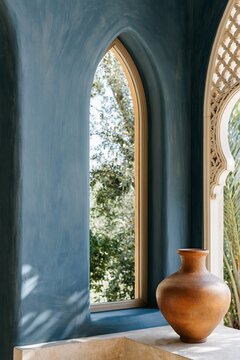 Arched window with deep indigo plaster wall and terracotta vessel on limestone sill, illuminated by warm Moroccan afternoon light