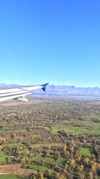 Airplane Window View of Mountains and Countryside. View from airplane window over mountains, lake and countryside during flight. Travel scene with aircraft wing, blue sky and aerial landscape.