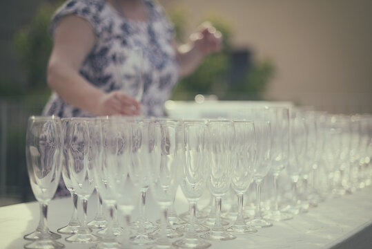 Rows of empty champagne flutes on a garden table with a woman in the background picking one up