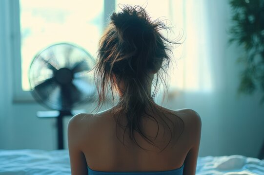 Back view of person with messy bun and bare shoulders sitting on bed facing a standing fan by a sunlit window, quiet contemplative morning mood