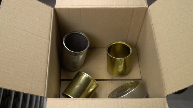 Different empty metal food cans are placed in a cardboard box, slow motion.  Disposal and recycling of metal containers