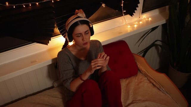 Top view mid shot of a young Caucasian woman wearing headphones, listening to music and staring blankly into the distance while scratching her hand with her fingers. Anxiety, nervous habits