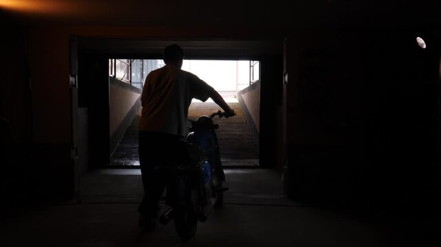 Man pushing his classic motorcycle up a concrete ramp, emerging from the dark underground garage into the bright daylight, creating a strong silhouette against the light