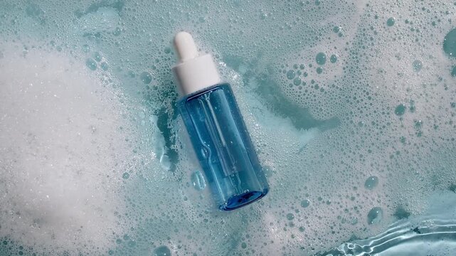 Bottle with a pipette for oil, toner, emulsion on water texture background surface with bubbles of white soapy foam. Women's cosmetics skincare, body care. Products advertisement spa, relaxation. 4k