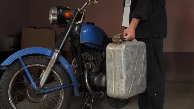 Person is standing in a garage, holding a retro metal petrol canister next to a classic blue motorcycle, preparing for a journey or maintenance, highlighting vintage transportation