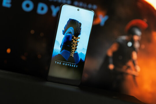 Screen displaying The Odyssey film poster by Christopher Nolan against blurred movie backdrop, cinema entertainment and mobile screen concept.