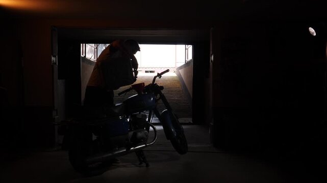 Man in silhouette bending over a vintage 1970s motorcycle, possibly refueling or performing maintenance in a dark garage or workshop entrance with bright sunlight in the background