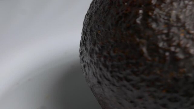 Close-up of a dark Hass avocado rotating on a white plate, showcasing its bumpy, textured skin and oval shape under bright studio lighting.