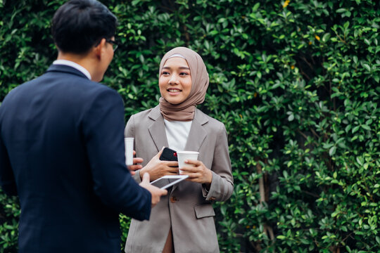Young business muslim woman and business man colleague walking and discussing outside the office. Woman is holding a coffee and businessman is holding a digital tablet outdoor.
