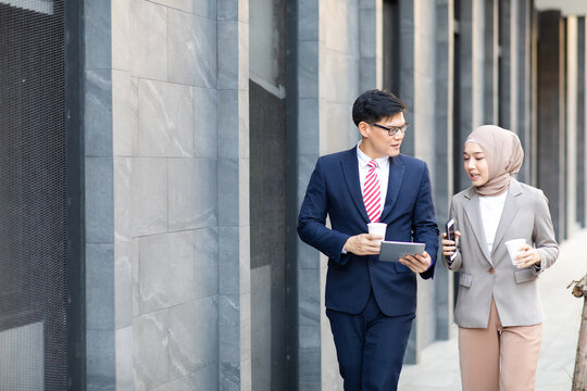 Young business muslim woman and business man colleague walking and discussing outside the office. Woman is holding a coffee and businessman is holding a digital tablet outdoor.