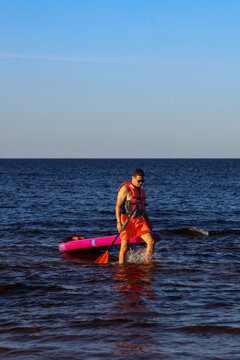 A man wearing a red life vest and orange shorts wades through choppy blue ocean water, holding a paddle with an orange blade.