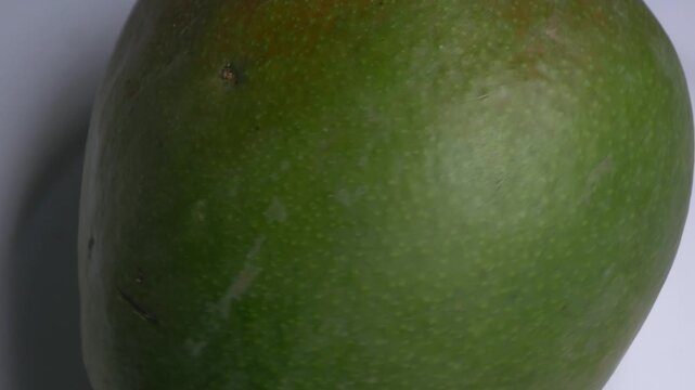 Close-up of a whole fresh green mango rotating on a white plate, showcasing its smooth skin, oval shape, and red blush under bright studio lighting.