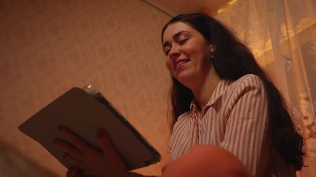 Young Caucasian woman with braces relaxing and smiling while watching an online video on a tablet in the evening. Digital entertainment, home leisure, dental care journey