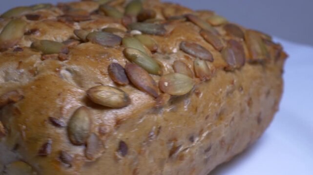 Close-up of a fresh whole grain bread roll with pumpkin seeds rotating on a white plate, showcasing its golden crust and textured surface under studio lights.