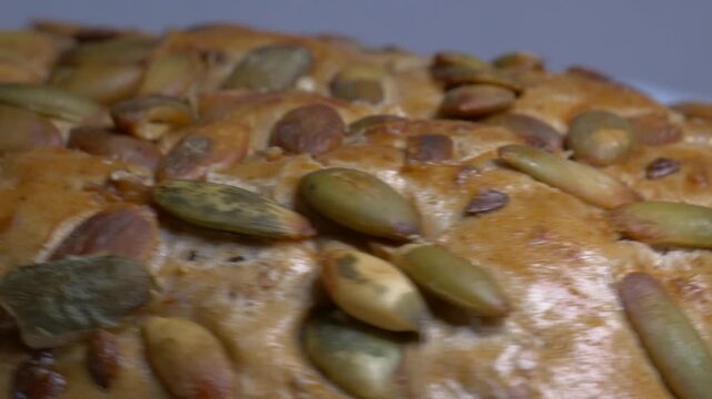 Close-up of a fresh whole grain bread roll with pumpkin seeds rotating on a white plate, showcasing its golden crust and textured surface under studio lights.