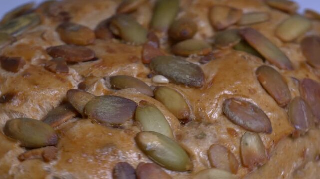 Close-up of a fresh whole grain bread roll with pumpkin seeds rotating on a white plate, showcasing its golden crust and textured surface under studio lights.