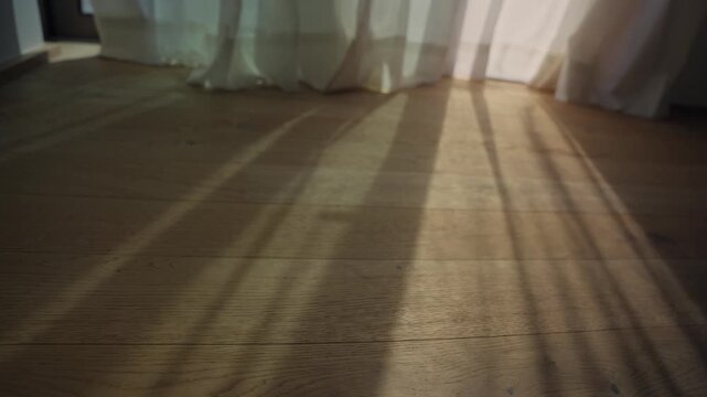 Rays of light from a window creating moving shadows on a parquet floor from dawn or morning. Sunlight shining inside house or apartment as curtain moves by slow cozy breeze of wind. Hotel room or home