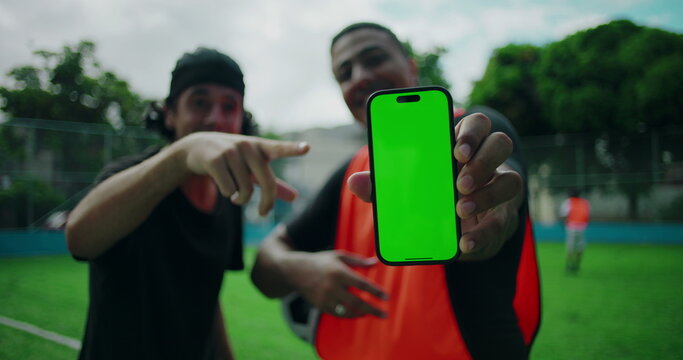 Two Hispanic men smiling and pointing at smartphone with green screen, presenting mobile app concept while standing on soccer field, expressing excitement