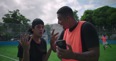 Two Hispanic men standing on soccer field, one showing shocking content on smartphone while friend reacts in disbelief, surprise and laughter, technology, friendship, outdoor recreation © Marco