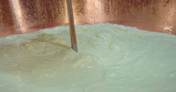 Slow Motion Of Mechanical stirring rod mixing fresh cheese curd in a traditional copper vat during dairy production. Cheese Curd Stirred inside Large Copper Dairy Vat.