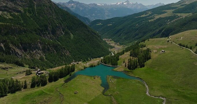 Drone flies toward a vibrant turquoise mountain reservoir fed by winding tributaries, revealing spectacular alpine peaks and pristine wilderness beneath a clear summer sky.